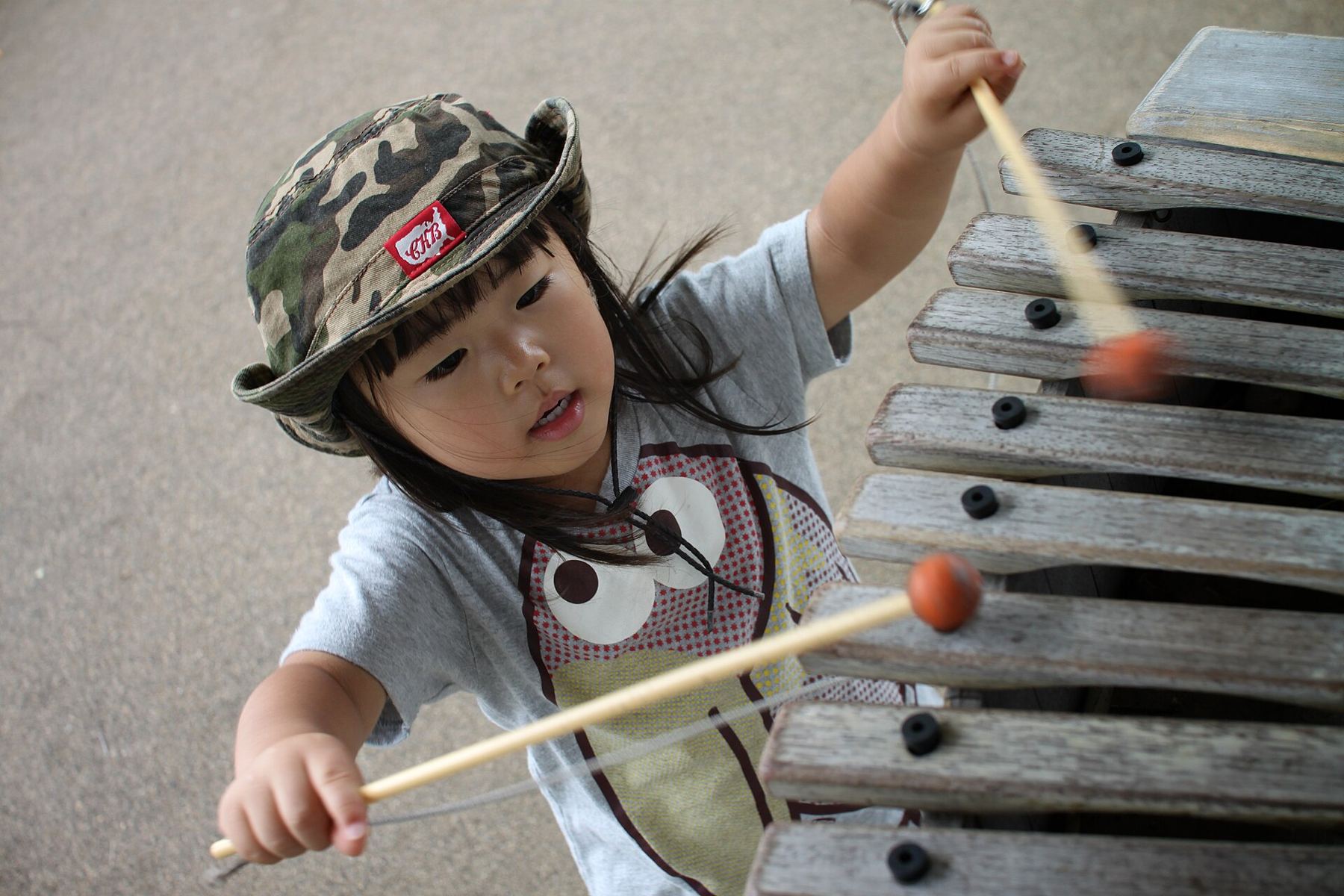 A young child in a sun hat playing a large xylophone with mallets
